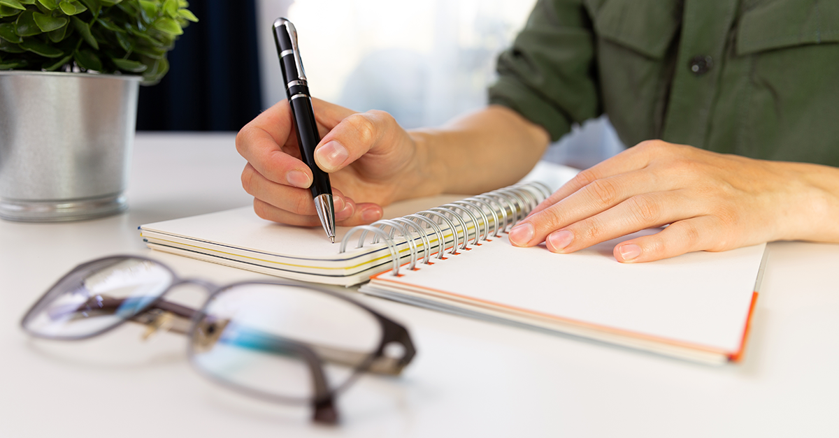 Person writing with a black pen in a spiral-bound notebook