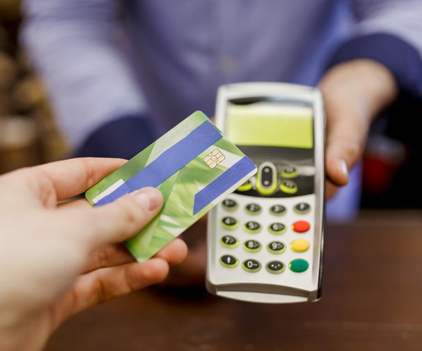 A customer's hand is seen holding a green credit card with blue stripes. The customer is paying at a mobile POS terminal held by an employee in a blue button down shirt. 